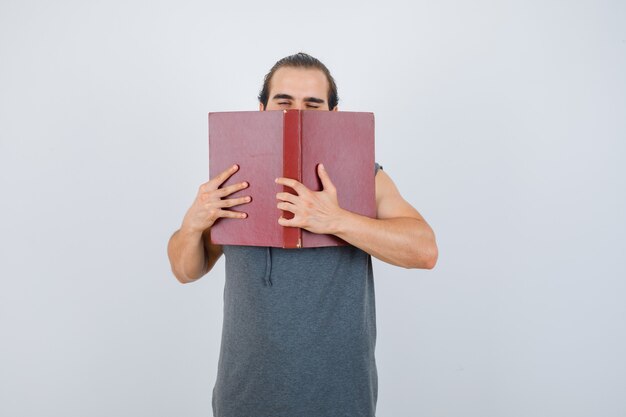 Varón joven en sudadera con capucha sin mangas que sostiene el libro abierto en la cara y mirando soñoliento, vista frontal.