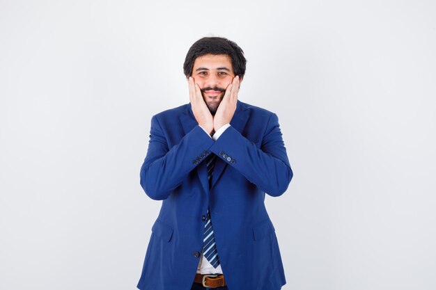 Varón joven tomados de la mano en la cara mientras sonríe en camisa, chaqueta, corbata y parece contento, vista frontal.