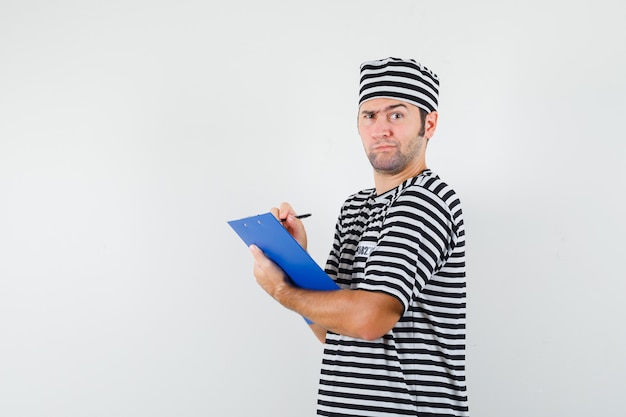 Varón joven tomando notas en el portapapeles en camiseta, sombrero y mirando pensativo.