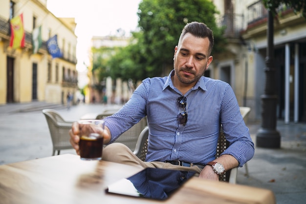 Varón joven en un traje formal sentado en un café al aire libre y bebiendo una bebida fría