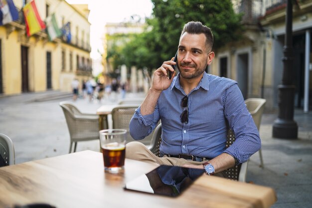 Varón joven en un traje formal sentado en un café al aire libre hablando por teléfono