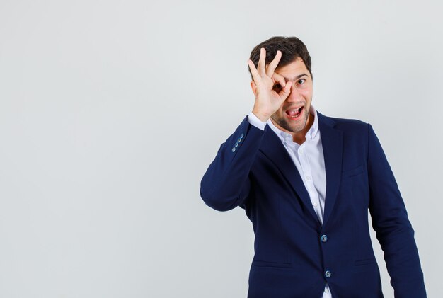 Varón joven en traje haciendo signo ok en los ojos y mirando alegre, vista frontal.