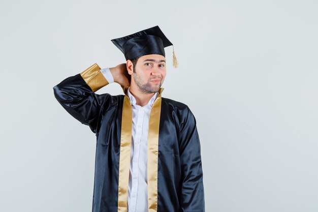 Varón joven en uniforme graduado sosteniendo la mano en el cuello y mirando elegante, vista frontal