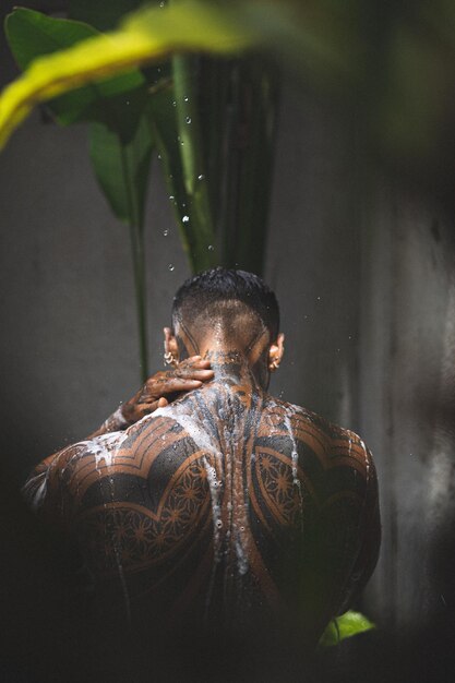 Varón latinoamericano musculoso sin camisa con tatuajes de pie tomando una ducha al aire libre después del entrenamiento en el fondo de hojas tropicales. Ducha en la villa privada.