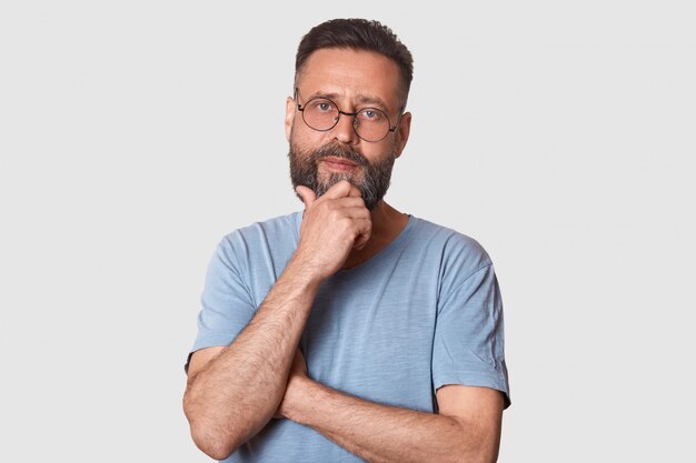 Varón de mediana edad, barbudo, con expresión facial pensativa, vestida con una camiseta informal gris y gafas redondas, mantiene la mano debajo de la barbilla, se ve pensativo, piensa en una nueva idea, tiene grandes planes.