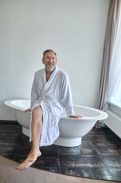 Varón relajado en una bata de baño de felpa blanca sentado con los pies descalzos en el borde del baño