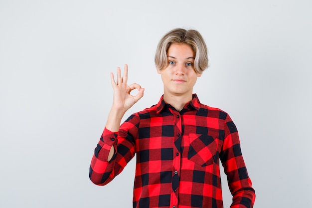 Varón rubio adolescente que muestra un gesto aceptable en camisa casual y que mira feliz, vista frontal.