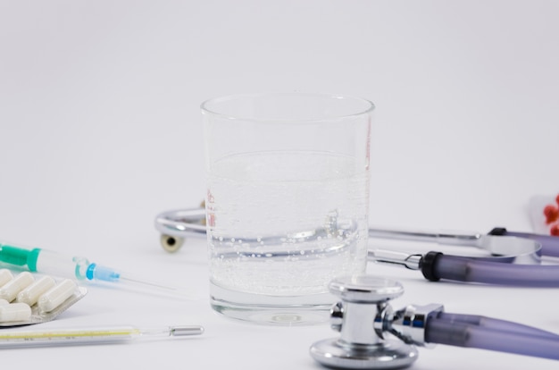 Vaso de agua; estetoscopio; pastillas Jeringa y termómetro sobre fondo gris