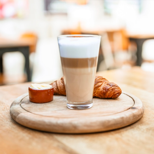 Vaso de café capuchino con croissant en bandeja de madera