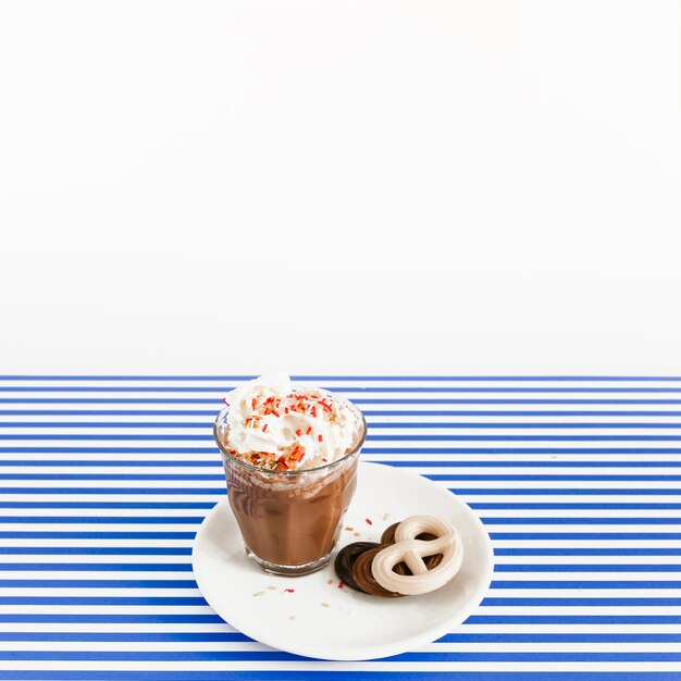 Vaso de café con crema batida y chocolates de pretzel en un plato sobre fondo de rayas blancas y azules