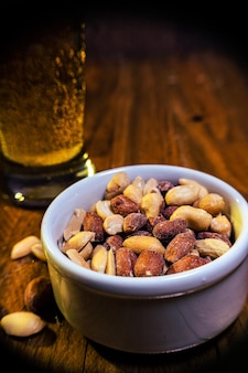 Vaso de cerveza lager con maní sobre la mesa y en un recipiente blanco sobre una mesa de madera.