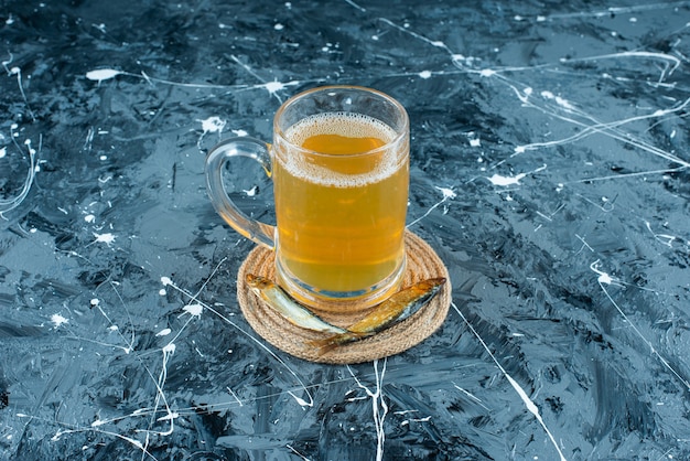 Un vaso de cerveza y pescado en un salvamanteles, sobre la mesa azul.