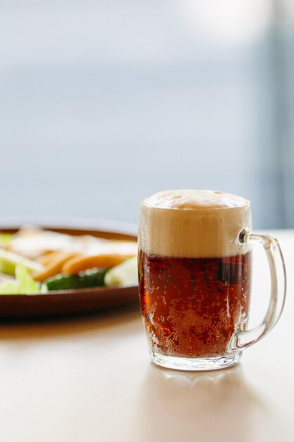 Vaso de cerveza de pie cerca de un plato de aperitivos Cerveza oscura con espuma alta y espesa De pie sobre una superficie de madera lisa de la mesa del restaurante o del pub Aspecto delicioso Frío y fresco Para compañía de amigos