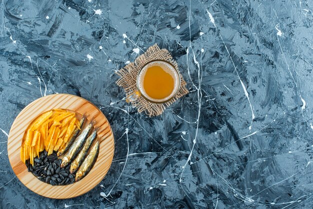 Un vaso de cerveza en textura y aperitivos en placa de madera, sobre la mesa azul.