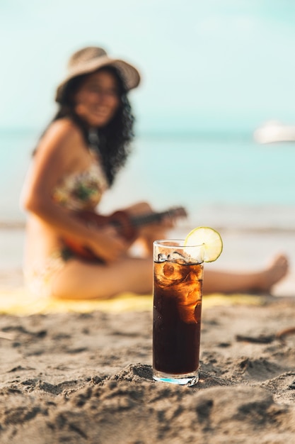 Vaso de coca cola con hielo y mujer en playa de arena