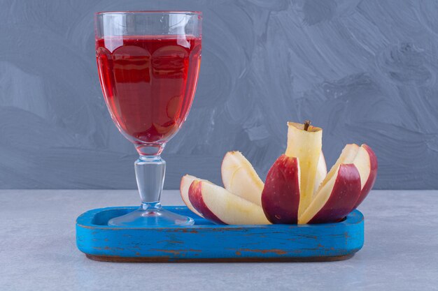 Un vaso de jugo de cereza y manzana en rodajas sobre una placa de madera sobre una mesa de mármol.