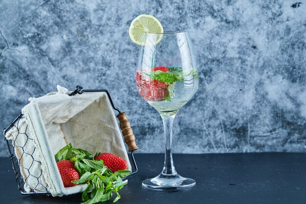 Un vaso de jugo con frutas enteras en el interior y una canasta de fresas en la superficie azul