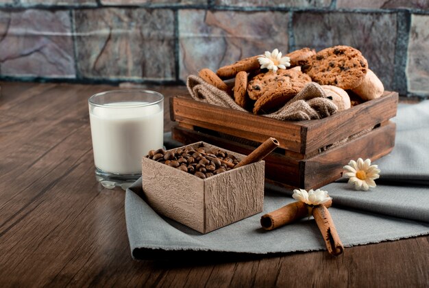 Un vaso de leche con bandeja de galletas y caja de granos de café sobre una mesa de madera