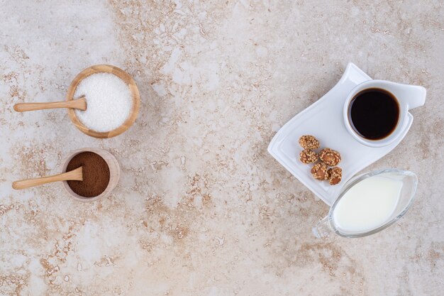 Un vaso de leche, tazones pequeños de azúcar y café molido en polvo, una taza de café y cacahuetes glaseados.