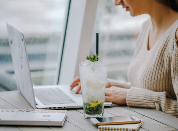 vaso de mojito junto a la mujer que trabaja en el cuaderno cerca de la ventana