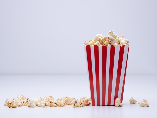 Vaso de papel de palomitas de maíz y algunas palomitas de maíz esparcidas sobre una superficie blanca