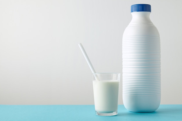 Vaso transparente con leche fresca y pajita blanca en el interior cerca de una botella de plástico en blanco con tapa azul