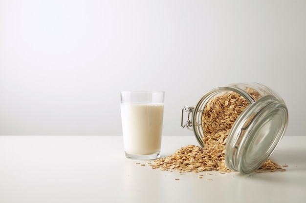 Foto gratuita vaso transparente con leche orgánica fresca cerca de un tarro rústico medio abierto con copos de avena esparcidos aislados en el centro en la vista lateral de la mesa blanca