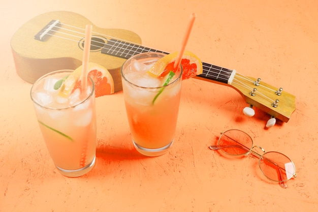 Vasos de jugo de cítricos con cubitos de hielo; Gafas de sol y ukelele en un telón de fondo con textura naranja