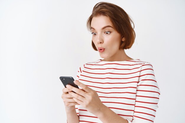 Vaya, mira esto. Chica sorprendida y emocionada mirando la pantalla del teléfono móvil, compras en línea con grandes descuentos, de pie en ropa casual contra la pared blanca