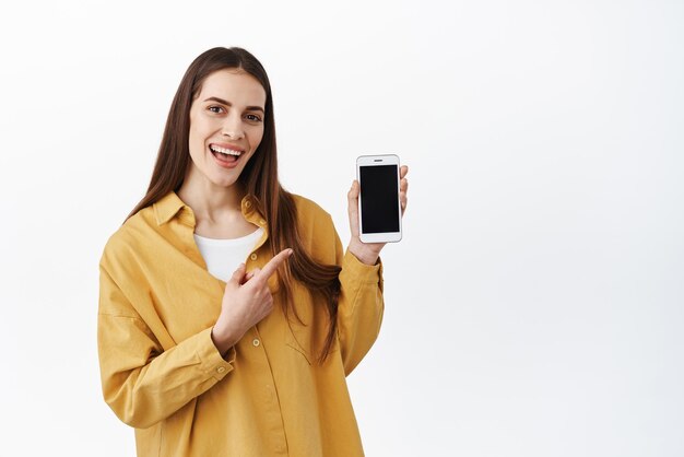Vaya a este sitio web Mujer joven sonriente señalando la pantalla en blanco del teléfono inteligente y anímelo a usar esta aplicación siga el enlace que muestra la aplicación de compras sobre fondo blanco