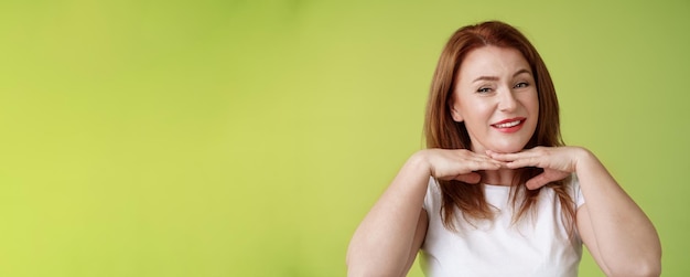 Foto gratuita se ve bien, feliz, alegre, pelirroja, mujer de mediana edad, sonriendo, encantada, tomándose de la mano bajo la barbilla.