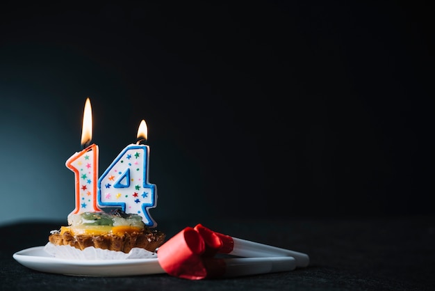 Vela encendida de cumpleaños número 14 en la rebanada de soplador de tartas y de fiesta contra el fondo negro