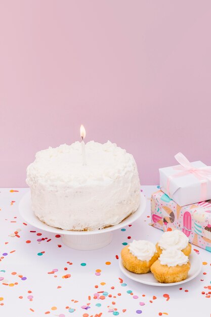 Vela encendida en pastel blanco con panecillos y regalos en escritorio blanco contra pared rosa