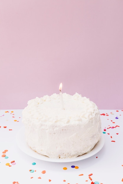 Una vela iluminada en la torta de cumpleaños blanca sobre la placa contra fondo rosado