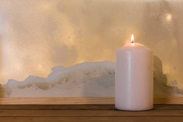 Vela en tablero de madera cerca de montón de nieve a través de la ventana