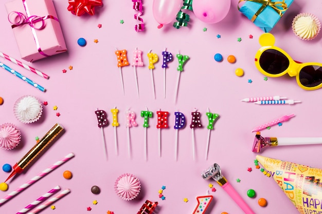 Velas de feliz cumpleaños rodeadas de serpentinas; gemas aalaw; Sombrero de fiesta y cuerno de fiesta sobre fondo rosa
