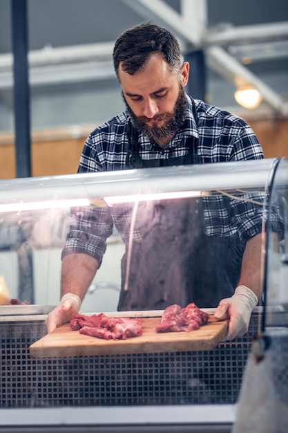 Vendedor de carne barbudo vestido con una camisa polar que sirve carne fresca cortada en un mercado.