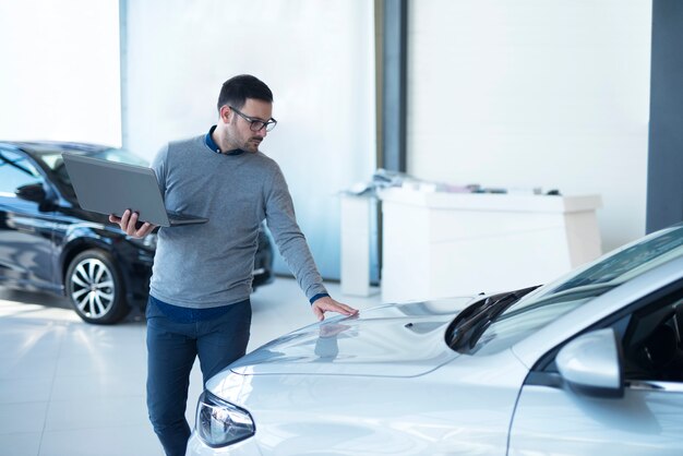 Vendedor de coches con portátil comprobando las especificaciones del vehículo en la sala de exposición del concesionario local