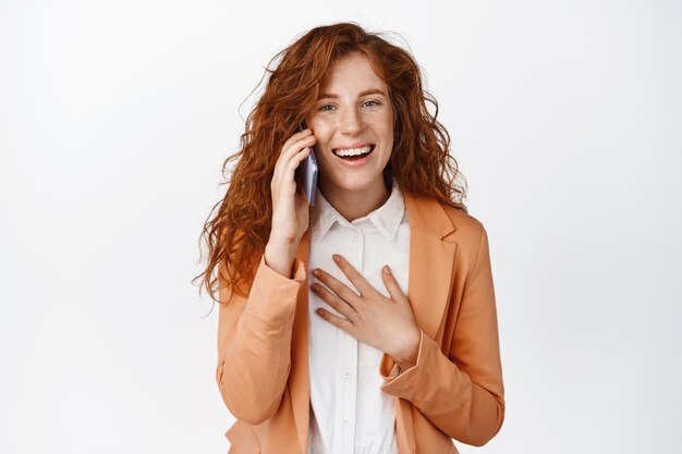 Vendedora alegre que tiene una llamada telefónica hablando por teléfono inteligente y riendo sonriendo divertida de pie sobre fondo blanco