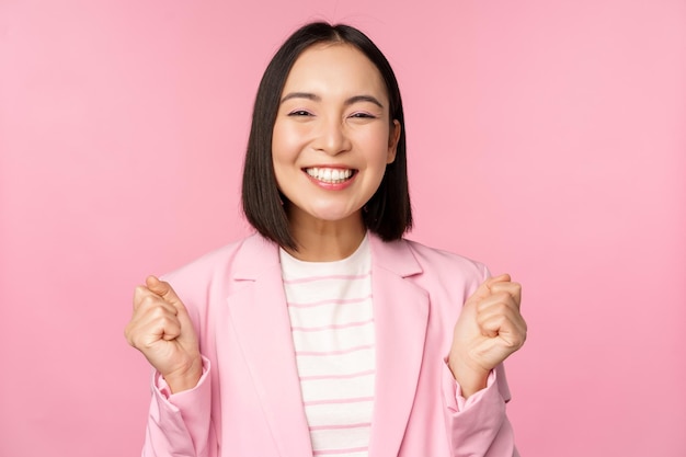 Una vendedora entusiasta, una mujer corporativa asiática, dice que sí, logra el objetivo y celebra el triunfo mirando con regocijo y sonriendo de pie sobre un fondo rosa