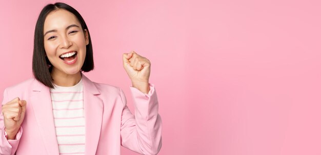 Una vendedora entusiasta, una mujer corporativa asiática, dice que sí, logra el objetivo y celebra el triunfo mirando con regocijo y sonriendo de pie sobre un fondo rosa