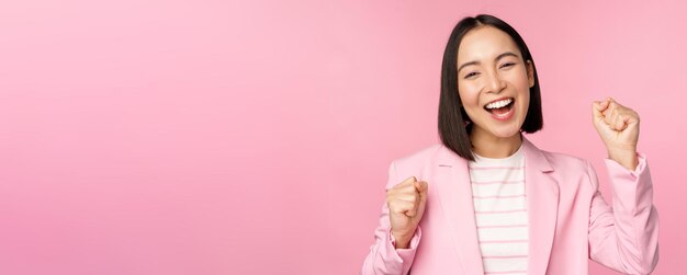 Una vendedora entusiasta, una mujer corporativa asiática, dice que sí, logra el objetivo y celebra el triunfo mirando con regocijo y sonriendo de pie sobre un fondo rosa