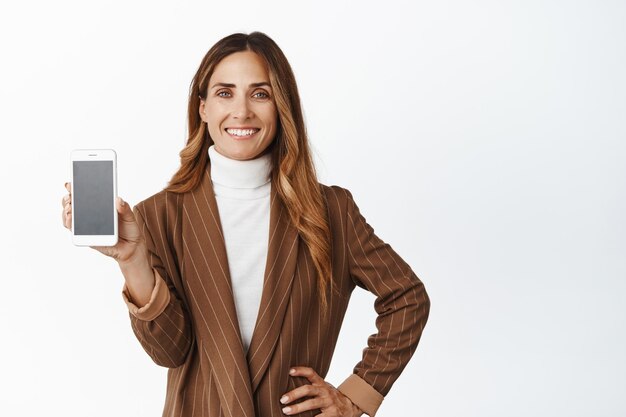 Vendedora segura de sí misma mujer de mediana edad que muestra la interfaz de la aplicación de pantalla vacía del teléfono inteligente de pie sobre fondo blanco