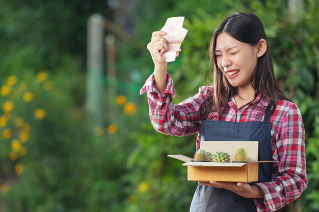 Venta de plantas en línea; La mujer se alegra mientras sostiene el dinero y la caja de envío llena con macetas de plantas