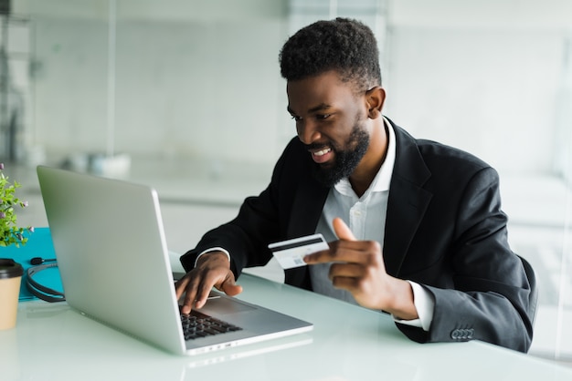 Ventas bancarias por internet. Exitoso empresario africano sentado en una computadora portátil y con tarjeta de crédito en la mano hasta que el empresario haga pedidos a través de Internet