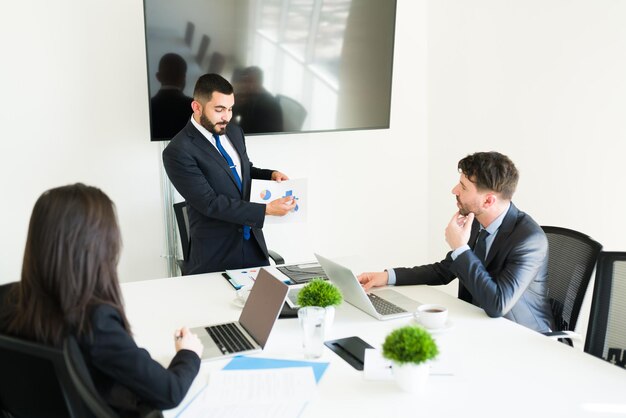 Las ventas están subiendo en la empresa. Atractivo representante de ventas que muestra un gráfico con los objetivos comerciales a sus compañeros de trabajo en la sala de reuniones