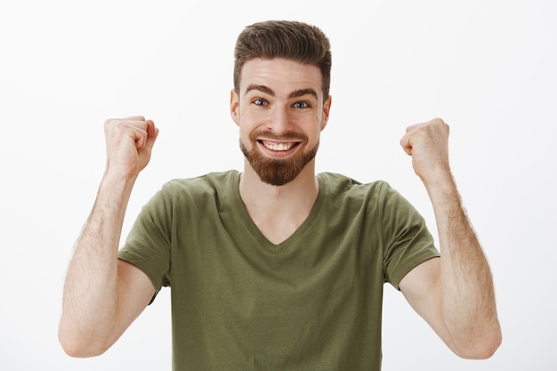 Ventilador masculino lindo activo alegre y enérgico con barba en camiseta levantando los puños apretados en la victoria y el triunfo celebrando la victoria del primer premio sonriendo de emoción y encantado con la pared blanca