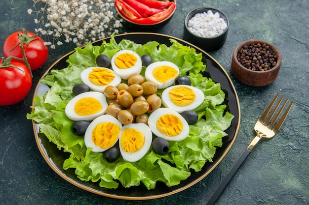 Ver arriba huevos cocidos en rodajas con ensalada verde y aceitunas sobre fondo azul oscuro