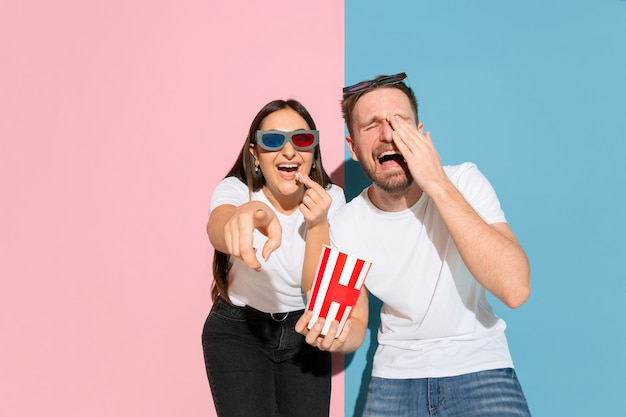 Ver cine en 3D con palomitas de maíz. Hombre y mujer joven y feliz en ropa casual en pared bicolor rosa, azul. Concepto de emociones humanas, expresión facial, relaciones, anuncio. Hermosa pareja.