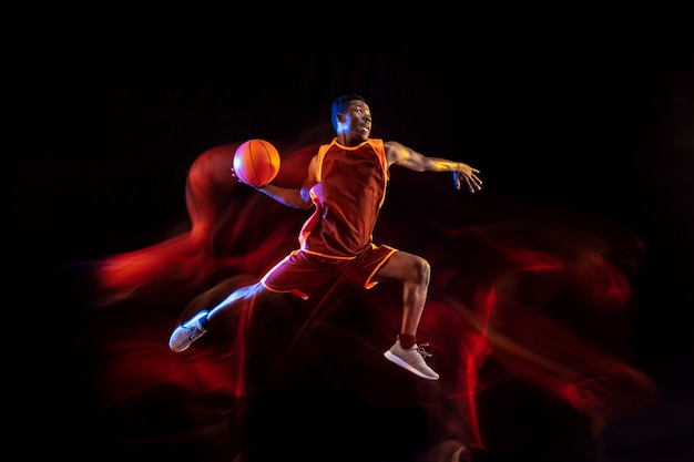 Ver el objetivo. Joven jugador de baloncesto afroamericano del equipo rojo en acción y luces de neón sobre fondo oscuro de estudio. Concepto de deporte, movimiento, energía y estilo de vida dinámico y saludable.
